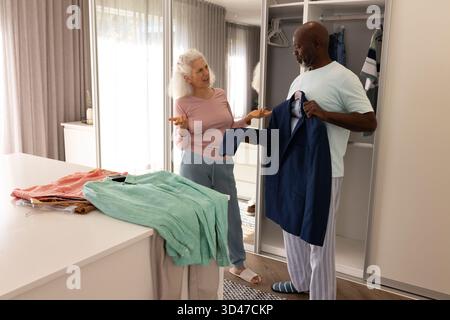 Über Outfit-Entscheidungen diskutieren Seniorenpaar, die sich für eine marineblaue Anzugsjacke im begehbaren Kleiderschrank mit Kleiderbügeln entscheiden Stockfoto