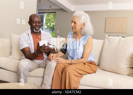 Diverse Seniorenpaare, die den Blutdruck auf dem Sofa im Wohnzimmer überprüfen, mit digitalem Monitor und Manschette Stockfoto