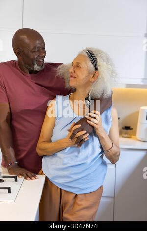 Ein vielseitiges Seniorenpaar, das sich in einer modernen Küche mit Airfryer und Kochfeld einen liebevollen Moment teilt Stockfoto