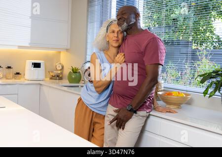 Liegendes Seniorenpaar, das sich in einer modernen weißen Küche umarmt, mit Airfryer und Obstschale Stockfoto