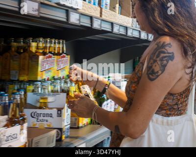 Cremona, Italien - 15. August 2025 Eine Frau mit einem großen anatomischen Herz-Tattoo auf dem linken Arm greift nach einer Flasche Corona Extra Bier aus einem Regal Stockfoto