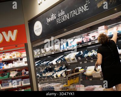 Cremona, Italien - 15. August 2025 Eine Frau mit lockigen braunen Haaren in einem schwarzen Kleid greift nach Produkten in einem Regal in einem hell beleuchteten Lebensmittelgeschäft Stockfoto
