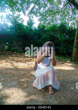 Frau mit farbenfrohem Schal und weißem Kleid, die sich in einer sonnendurchfluteten Outdoor-Umgebung hockt und ein Foto mit einer Vintage-Kamera macht Stockfoto