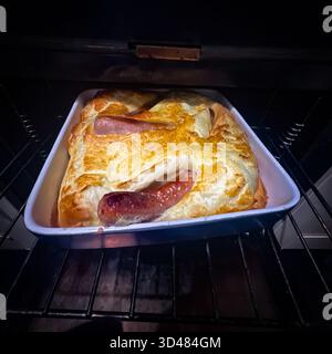 Eine Kröte im Loch wird aus dem Ofen geholt. Kröte in the Hole ist ein klassisches britisches Komfortgericht: Würstchen, gebacken in einem leichten, luftigen Yorkshire-Pudding Stockfoto