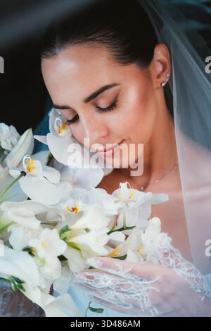 Bride hält sanft einen Blumenstrauß weißer Orchideen mit geschlossenen Augen und umhüllt den Moment mit einem ruhigen Ausdruck. Ihre Spitzenhandschuhe und der Schleier verleihen ihr einen Hauch von Ele Stockfoto