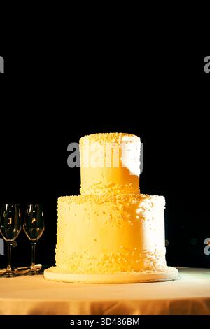 Zweistöckige Hochzeitstorte, elegant mit Perlen dekoriert, auf einem Tisch mit drei Weingläsern daneben, vor dunklem Hintergrund, wodurch ein Stockfoto