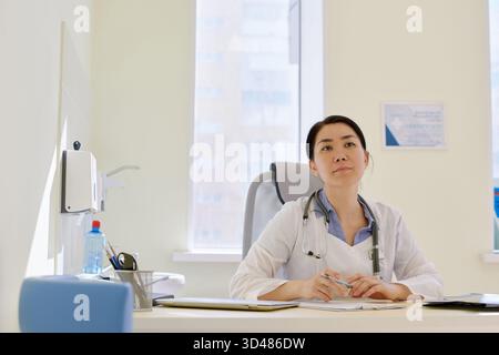 Junge asiatische Ärztin mit Stethoskop am Arbeitstisch im Sprechzimmer sitzend und in die Ferne blickend Stockfoto
