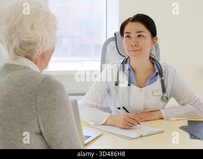Ältere Frau mit ärztlicher Beratung im Krankenhaus. Junge asiatische Ärztin, die dem Seniorinnen aufmerksam zuhört und freundlich lächelt Stockfoto