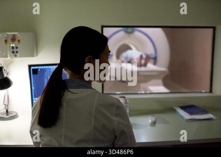 Rückansicht einer Ärztin, die das Bild am Computer überprüft, während ihre Patientin im modernen Krankenhaus MRT-Scan durchläuft Stockfoto
