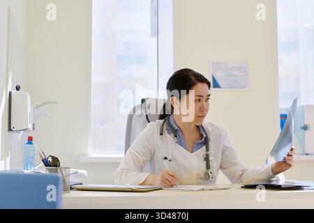 Professionelle Radiologin sitzt am Tisch im Sprechzimmer und untersucht Röntgenaufnahmen von Patienten nach MRT-Verfahren Stockfoto