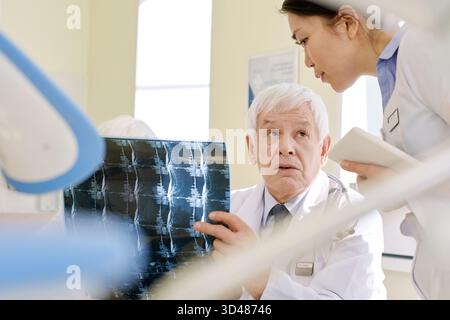 Reifer männlicher Radiologe bei Röntgenuntersuchung des Patienten nach MRT-Verfahren und Beratung mit junger weiblicher Assistentin in der Klinik Stockfoto