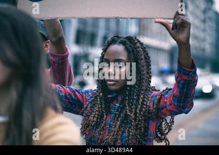 Eine junge Frau mit lockigen Haaren steht fest in einer Stadtstraße und hält ein Schild über dem Kopf. Die Menschen um sie herum sind in einem lebhaften Protest verwickelt, Emphas Stockfoto