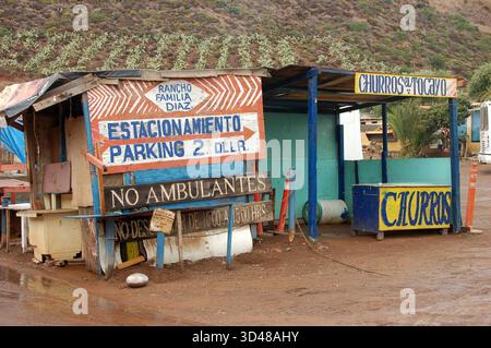 Kleine rustikale Parkplätze und Churros stehen in Ensenada, Mexiko, mit bunten handgefertigten Schildern auf einer Feldstraße und Hügeln im Hintergrund Stockfoto