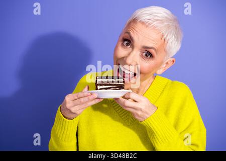 Eine ältere Frau mit kurzen weißen Haaren lächelt, während sie ein Stück Schokoladenkuchen auf einem Teller vor einem leuchtend violetten Hintergrund genießt Stockfoto
