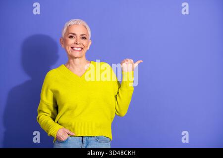 Hell gekleidete ältere Frau lächelt und zeigt vor blauem Hintergrund mit einem gelben Pullover, der einen fröhlichen Lebensstil festlegt Stockfoto
