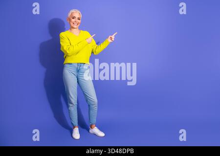 Elegante, reife Frau in einem leuchtend gelben Pullover zeigt mit Freude vor einem lila Hintergrund, der lässige Mode und Wohlbefinden zeigt Stockfoto