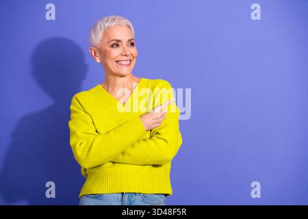 Eine ältere Frau mit kurzen weißen Haaren in Pullover lächelt und zeigt nach rechts vor einem violetten Studio-Hintergrund für Lifestyle-Mode Stockfoto