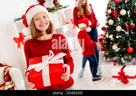 Lächelndes Mädchen in rotem Pullover Weihnachtsmütze präsentiert ein Geschenk in einem gemütlichen Zuhause während Weihnachten, während eine Familie sich in der Nähe des Baumes versammelt Stockfoto