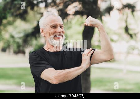 Senior Mann im Park im Freien lächelt, während er seinen Arm in einer Fitnessposition beugt und an einem sonnigen Tag einen gesunden aktiven Lebensstil an der frischen Luft genießt Stockfoto