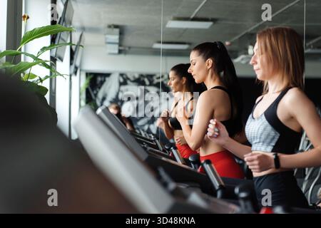 Eine Gruppe von zwei schlanken aktiven Frauen in Sportbekleidung, die auf dem Laufband im Fitnessclub Herz-Kreislauf-Übungen machen. Freundinnen mit dunklem Haar trainieren gemeinsam im modernen G Stockfoto