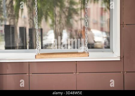 Holzschaukel, die an einem reflektierten Glasfenster mit urbanem Hintergrund hängt. Stockfoto