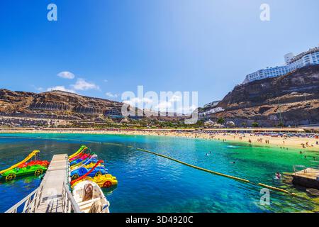 Sonniger Blick auf den Strand mit Touristen, die im Atlantischen Ozean schwimmen, und farbenfrohen Tretbooten, die in der Nähe des Piers auf dem Hintergrund des RIU Hotels geparkt sind. Grand Canary. Spanien Stockfoto