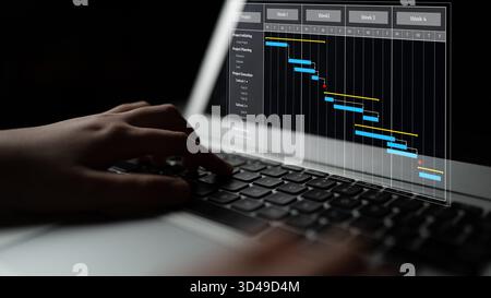 Eine fokussierte Ansicht der Hände, die auf einer Laptop-Tastatur tippen, mit einem Gantt-Diagramm auf dem Bildschirm. Dieses Bild stellt Themen des Projektmanagements dar Stockfoto