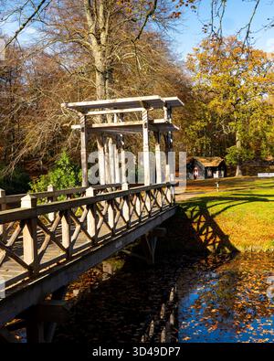 Holzbrücke über einen ruhigen Teich, umgeben von Herbstbäumen im warmen Sonnenlicht. Malerischer Blick auf ein Holzgebäude in einem friedlichen Park im Herbst. Stockfoto
