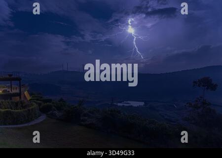 Eine dramatische Nachtszene in den Bergen von Sri Lanka, die einen starken Blitz einfängt, der die Landschaft und entfernte Windräder beleuchtet. Stockfoto