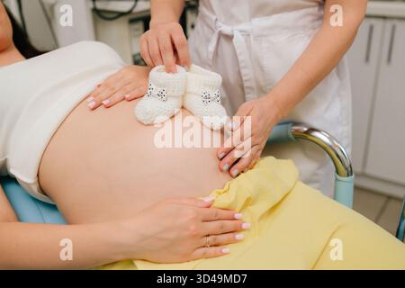 Schwangere Frau und Gynäkologin mit einem Paar gestrickter Babyschuhe auf dem Bauch. Ich erwarte Mutter mit kleinen Babyschuhen, während ich auf ihr Baby wartete Stockfoto