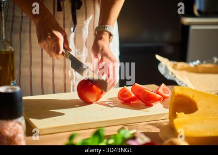 Nahaufnahme einer Frau, die Tomaten auf Holzbrett in der Küche schneidet, mit Gemüse und Kürbis auf dem Tisch. Konzept von Bio-Lebensmitteln und gesundem Kochen Stockfoto