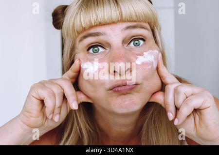 Attraktive Blondine mit blauäugigen Augen mittleren Alters, mit gebundenen Pferdeschwänzen auf den Haaren, sieht im Badezimmer im Spiegel, trägt Creme gegen Falten, Altersflecken auf, wh Stockfoto