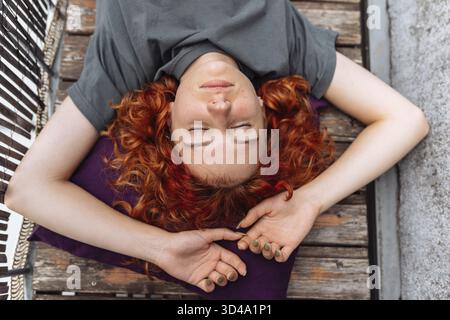 Porträt rothaarige lockige junge Frau, die sich auf Balkon oder Terrasse entspannen kann. Rothaarige, lockige junge Frau, die auf Holzdielen auf dem Balkon liegt Stockfoto
