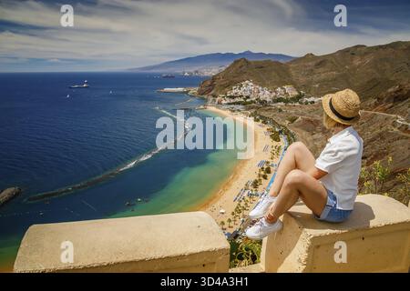 Eine Touristenfrau genießt einen Panoramablick auf den wunderschönen Strand Las Teresitas auf Teneriffa, umgeben von Palmen und einer gewundenen Straße Stockfoto