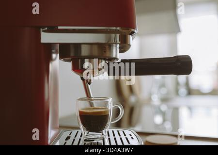 Espresso, der in der Küche in eine transparente Tasse aus der Kaffeemaschine gegossen wird, vor verschwommenem Hintergrundlicht Stockfoto