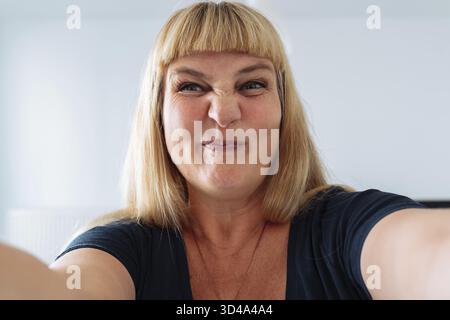Blauäugige attraktive positive Frau mittleren Alters, blond, grimaces, lächelt, zieht Aufmerksamkeit auf sich, in den Spiegel schauen, Selfie machen. Dating via Internet. Stockfoto
