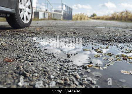 Pfützen Sie sich in einem Loch auf einer Landstraße mit Schotter, mit Reflektionen des Himmels, gefallenen gelben Blättern und einem Autorad. Herbstdepressive Realität. Straßenreparatur Stockfoto