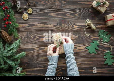 Flache rustikale Weihnachtskomposition mit Händen, die eine kleine Geschenkbox dekorieren, mit Tannenzweigen und Weihnachtsdekorationen auf braunem Holzhintergrund Stockfoto