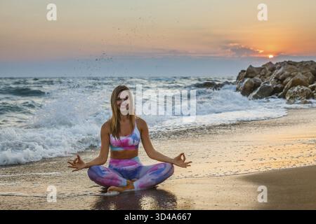 Lächelnde Frau, die Yoga praktiziert, sitzend in Lotusposition am Strand während Sonnenuntergang. Innere Harmonie und Körperpflege Stockfoto