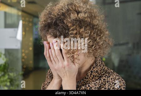 Geschweifte junge Frau mit trauriger Ausdrucksweise, die beim Schreien im Amt das Gesicht mit den Händen bedeckt Stockfoto