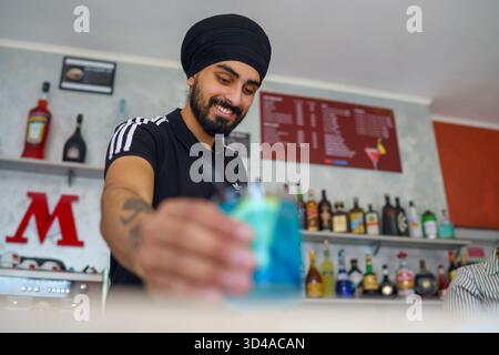 Crema, Italien - 25. August 2025 lächelnder Barkeeper mit Turban serviert einen lebhaften blauen Cocktail hinter einer Bar mit alkoholischen Getränken und Menü Boa Stockfoto