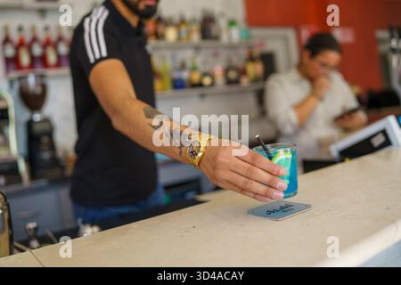 Crema, Italien - 25. August 2025 der Barkeeper platziert einen farbenfrohen tropischen Cocktail mit Eis und Garnelen an der Bar, während ein Kollege im Hintergrund arbeitet Stockfoto