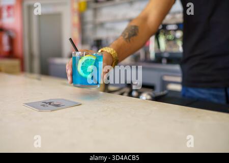 Crema, Italien - am 25. August 2025 serviert der Bartender einen erfrischenden blauen Cocktail mit Limettenscheibe und schwarzem Strohhalm auf einer Arbeitsplatte Stockfoto
