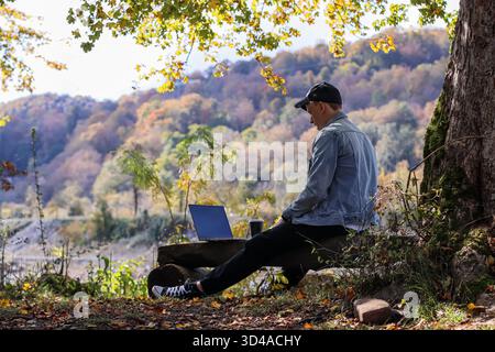Rückansicht eines Mannes, der im Herbstwald einen Laptop auf einem Holzstamm mit Kaffee benutzt. Von hinten sitzt ein Mann in Denim auf Holz, arbeitet am Laptop, eine Kaffeetasse ist sichtbar Stockfoto