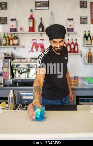 Crema, Italien - 25. August 2025 junger Sikh-Barkeeper mit schwarzem Turban und Bart, der fachmännisch einen leuchtenden blauen Cocktail an einer modernen Bar zubereitete Stockfoto
