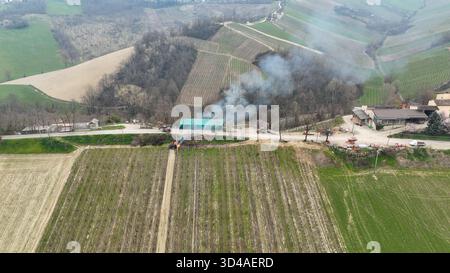 Aus der Vogelperspektive brennende Weinbauzweige, die unter einem bewölkten Himmel Rauch zwischen den rollenden Piacenza-Hügeln in Norditalien erzeugen Stockfoto