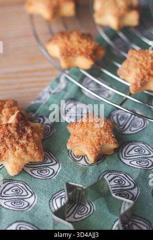 Frisch gebackene Zimt-Sternkekse auf einem Kühlregal, mit einem Metall-Stern-Ausstecher im Vordergrund. Gemütliche hausgemachte Weihnachtsbacken. Stockfoto