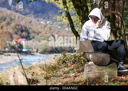 Junger Mann im Kapuzenpullover hält im Herbst das Telefon an das Ohr mit dem Laptop. Junger Mann in weißem Hoodie spricht auf Handy neben offenem Laptop Stockfoto