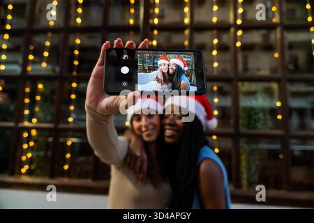 Verschiedene Frauen mit weihnachtsmützen machen während der weihnachtsfeier ein fröhliches Selfie auf einem Smartphone Stockfoto