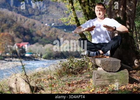 Mann im Kreuz auf Baumstämmen in der Nähe von Bäumen, Arme ausstrecken, Palmen offen, die auf Fluss und Berglandschaft gerichtet sind. Erwachsene Männer in Freizeitkleidung Stockfoto